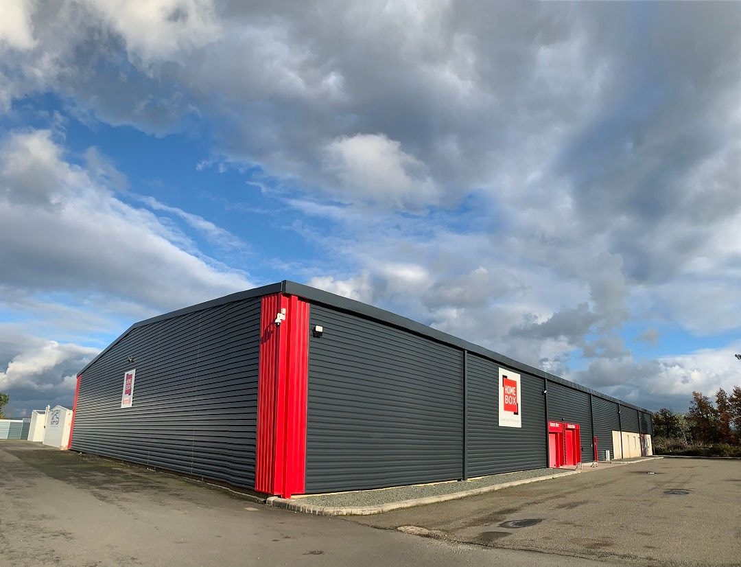 Dark grey warehouse with red accents under a cloudy sky and empty parking lot at HOMEBOX Le Mans, 95 route du chêne, Arnage