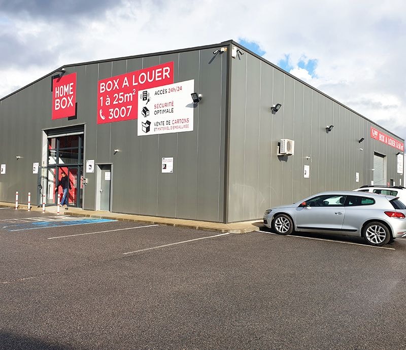 Grey metal building with red HOMEBOX panel and parked car under cloudy sky at HOMEBOX Ajaccio, RN 194 Baleone, Mezzavia