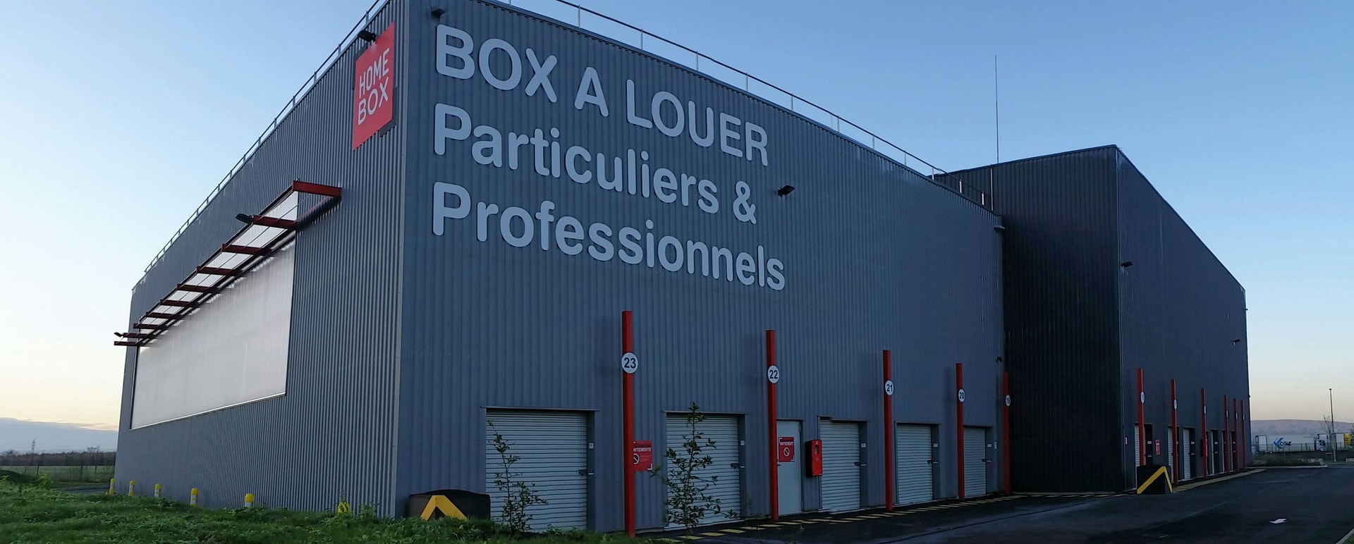 Grey corrugated metal warehouse with large box rental signage and red loading bay doors at HOMEBOX Carrières-Chanteloup, 5 rue Henri Navier, Chanteloup-Les-Vignes