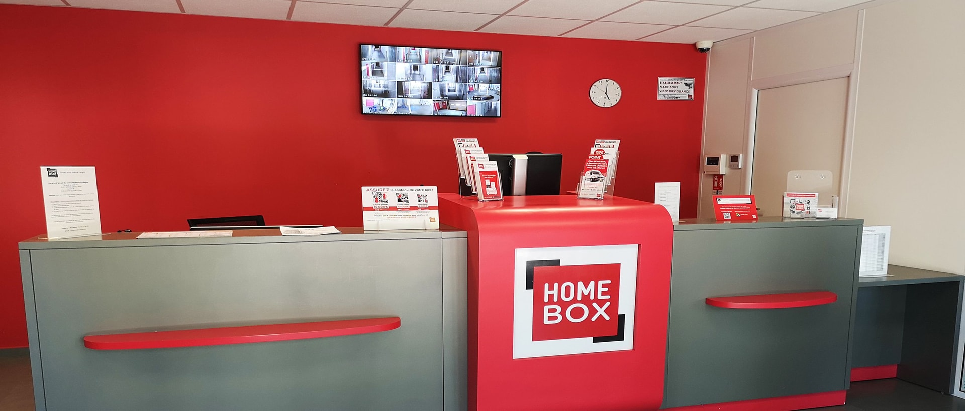 Red reception desk with HOMEBOX logo, wall-mounted surveillance screen and counter brochures at HOMEBOX Paris Ternes