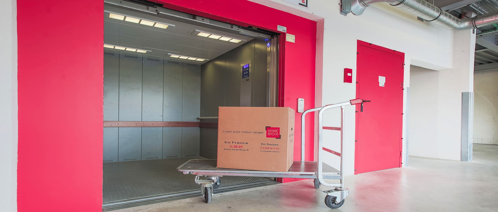 Cardboard box on trolley in front of large freight elevator with red walls at HOMEBOX Vannes Nord