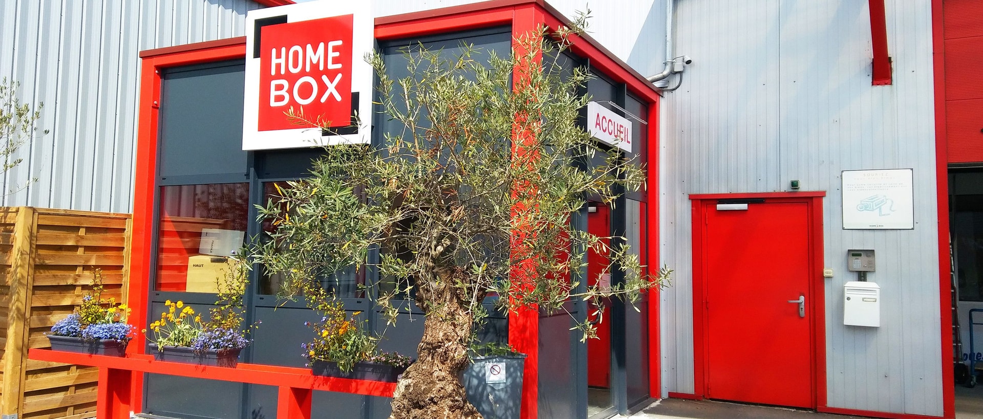 Red and grey entrance with olive tree, signage and Accueil door at HOMEBOX Caen Ouest, 5 rue des Vallees, Bretteville Sur Odon