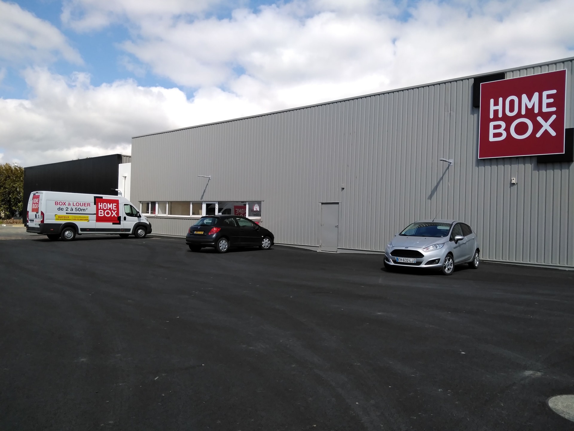 Grey warehouse facade with red HOMEBOX sign and branded van in parking lot at HOMEBOX Bayeux, 5 rue de la Résistance, Bayeux