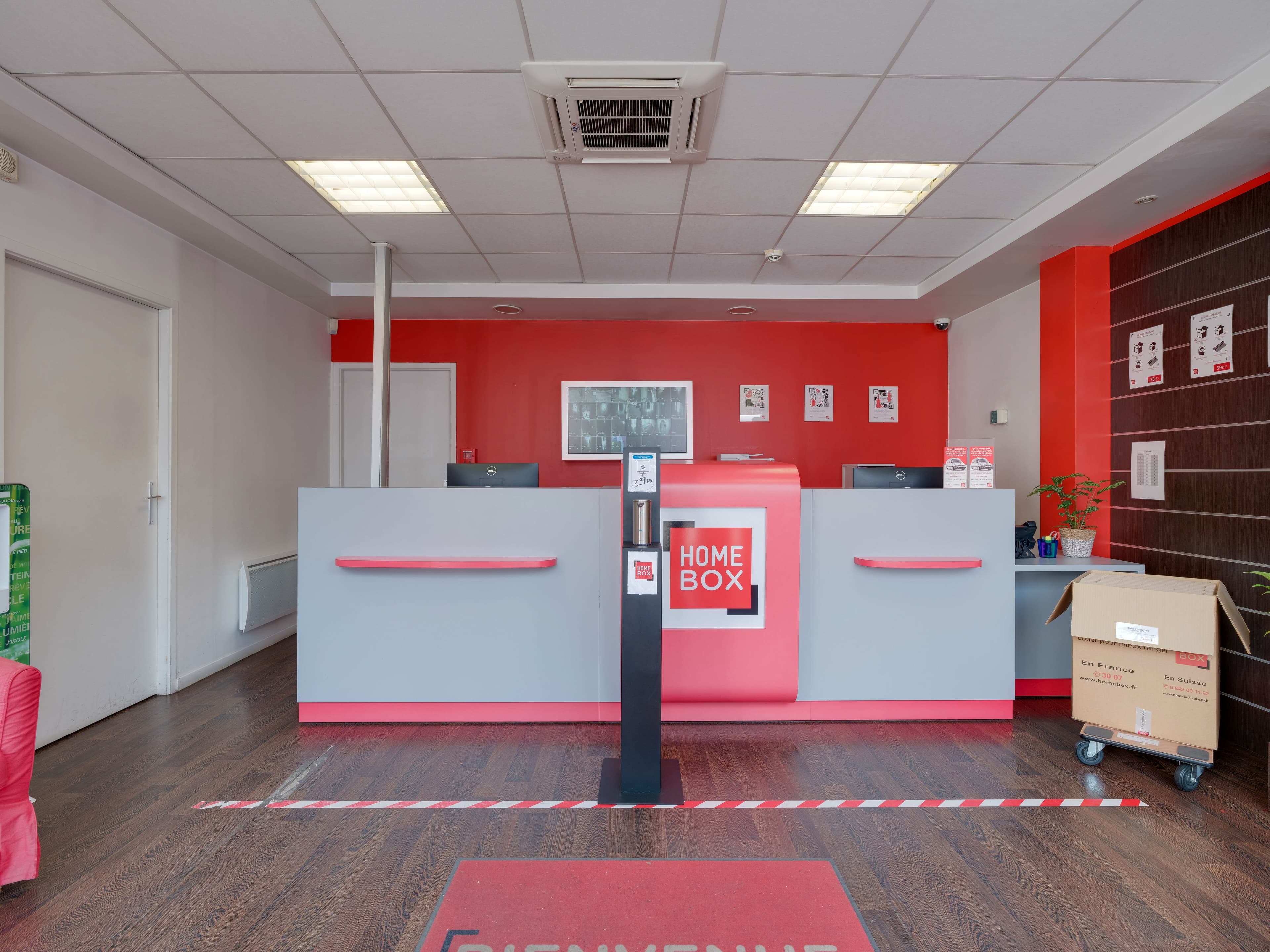 Comptoir d’accueil du centre HOMEBOX Annecy avec mur rouge et écran mural.
