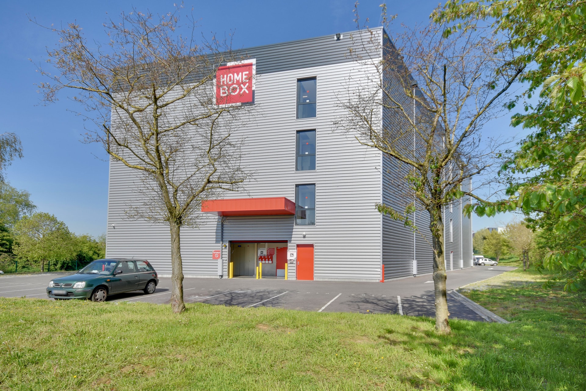 Façade bardage gris avec enseigne et auvent rouge vue depuis le parking, HOMEBOX Lille Lomme, Rue du Traité de Rome à Lomme