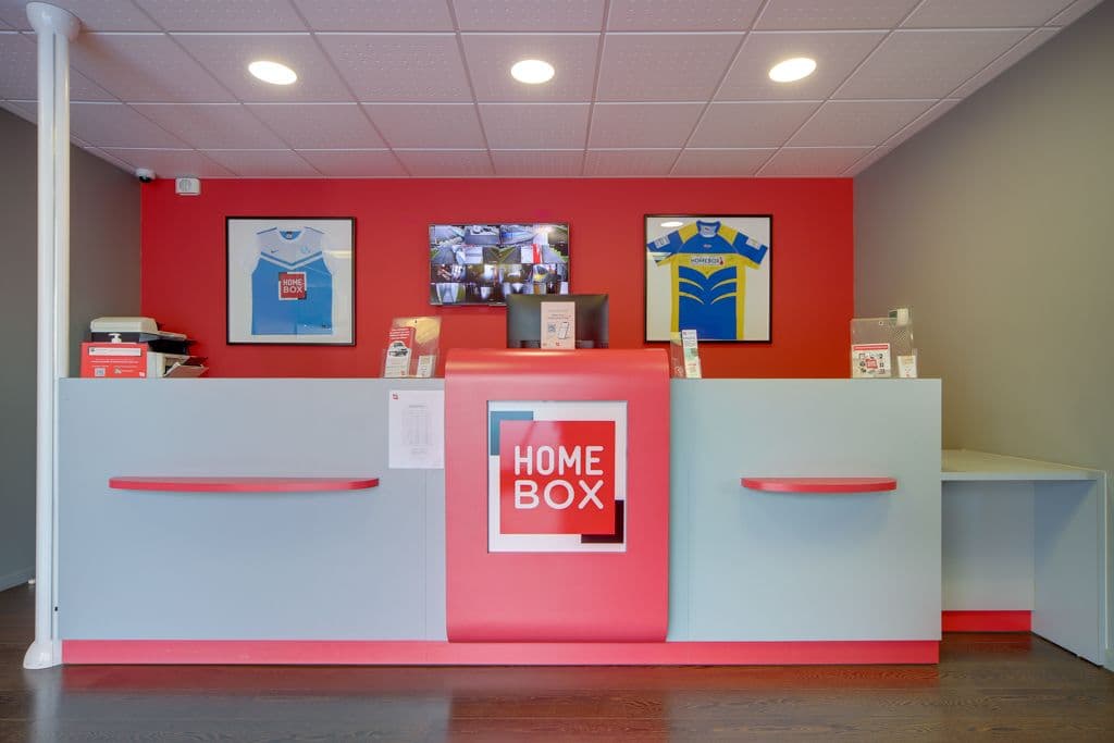 Accueil du centre HOMEBOX Blois avec comptoir rouge et gris et écrans de vidéosurveillance.