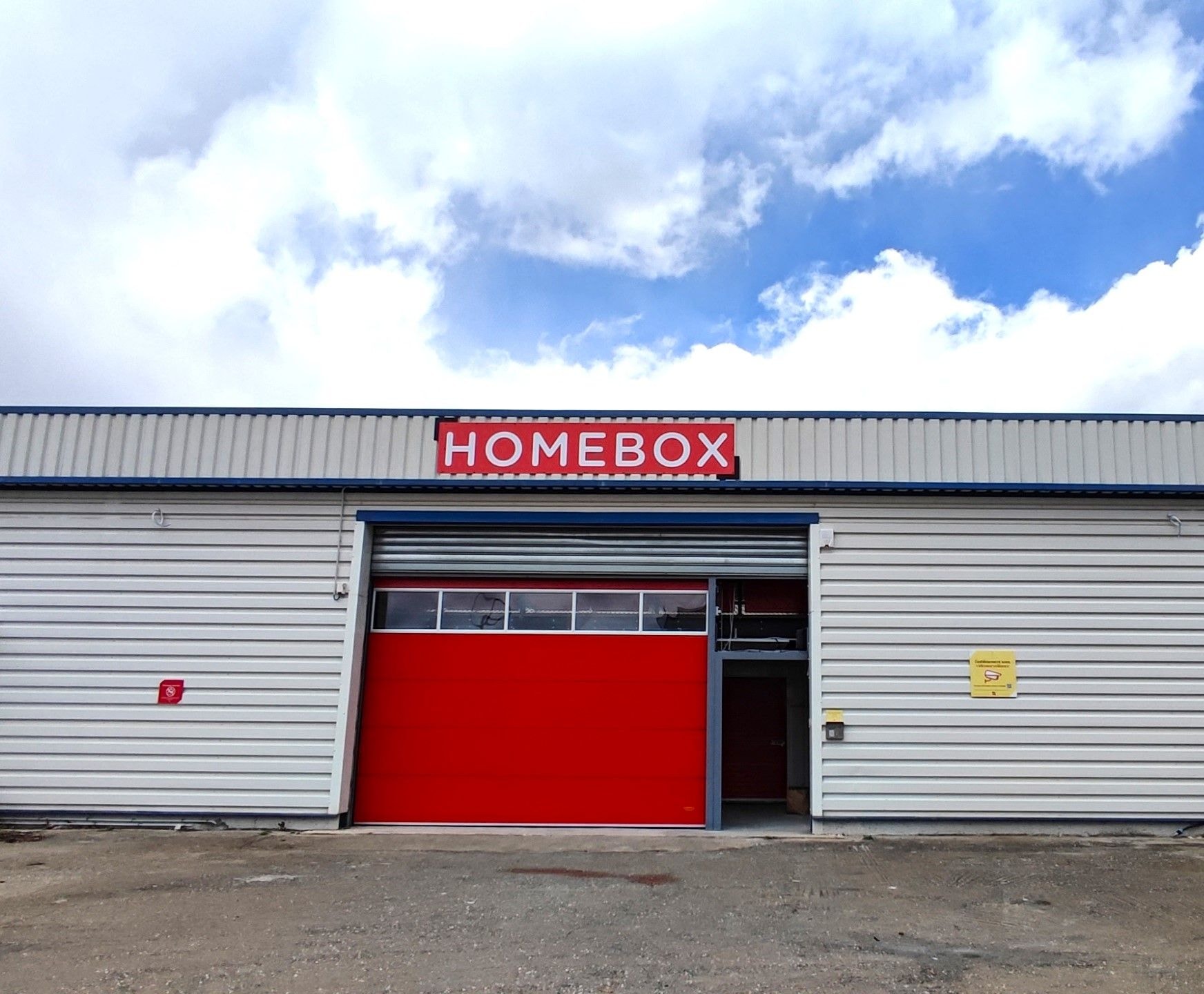 White warehouse with red rolling door and HOMEBOX sign under blue sky at HOMEBOX Corte, 19C Zone d'activité artisanale, CORTE