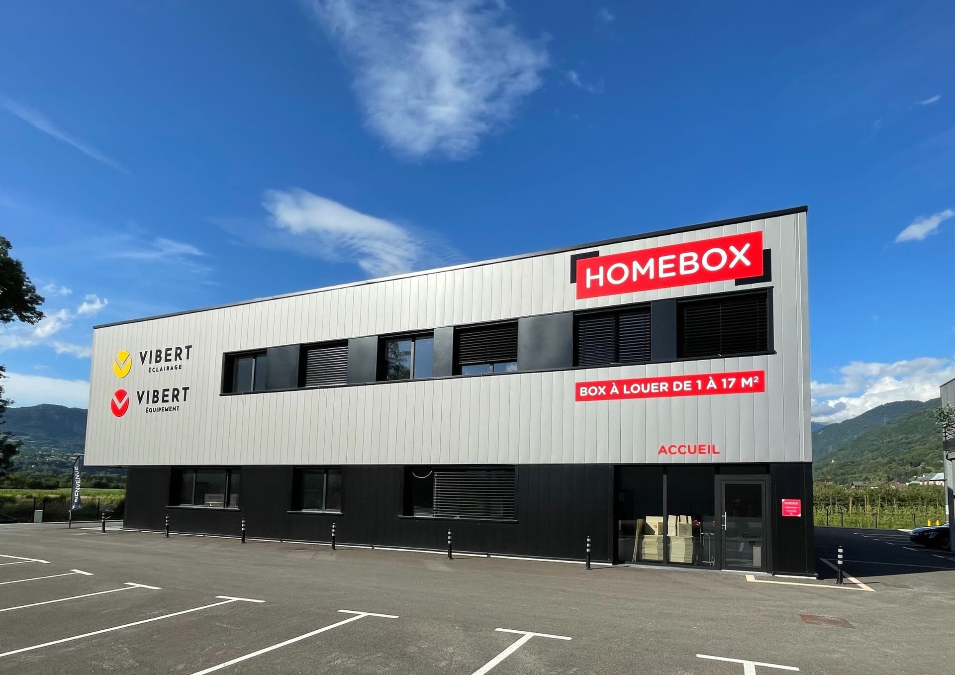 Modern black and white facade with red HOMEBOX sign and empty parking lot at HOMEBOX Albertville, ZAC Du Rotey, Notre-Dame-des-Millières