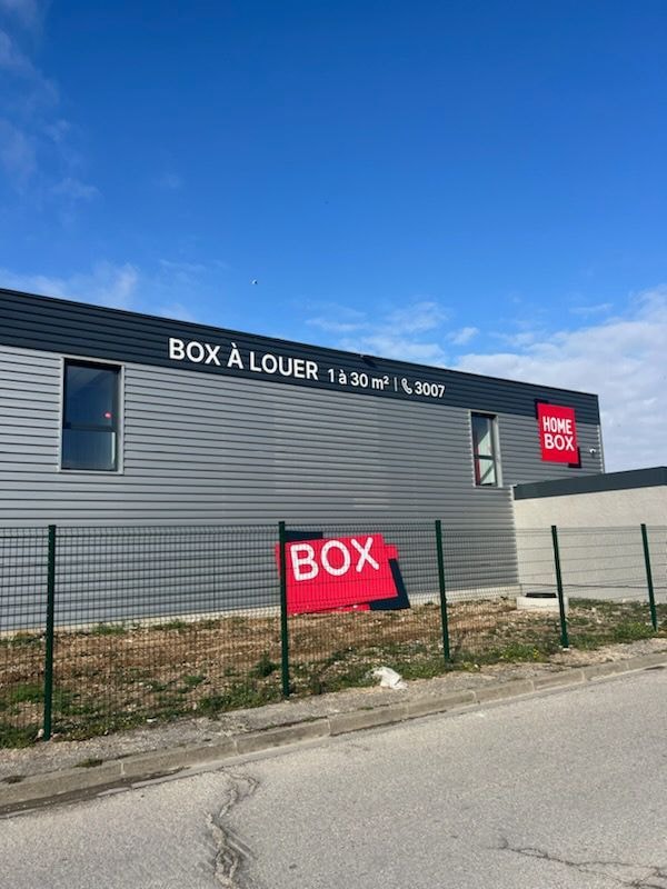 Grey facade with Box A Louer signage and red HOMEBOX logo behind fenced sidewalk at HOMEBOX Vitrolles, 1 rue de la Glacière ZI LES BAGNOLS, Vitrolles